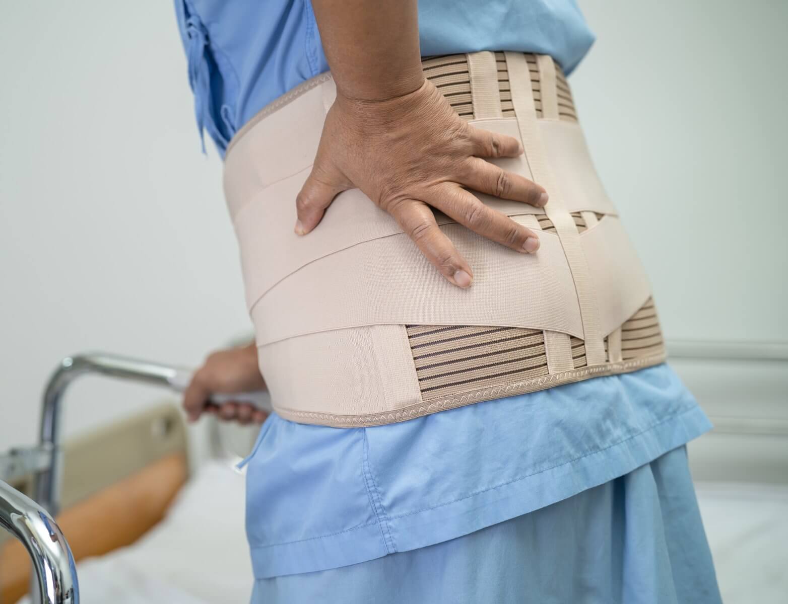 Patient in a medical setting wearing a back support brace, gripping a walker for support.